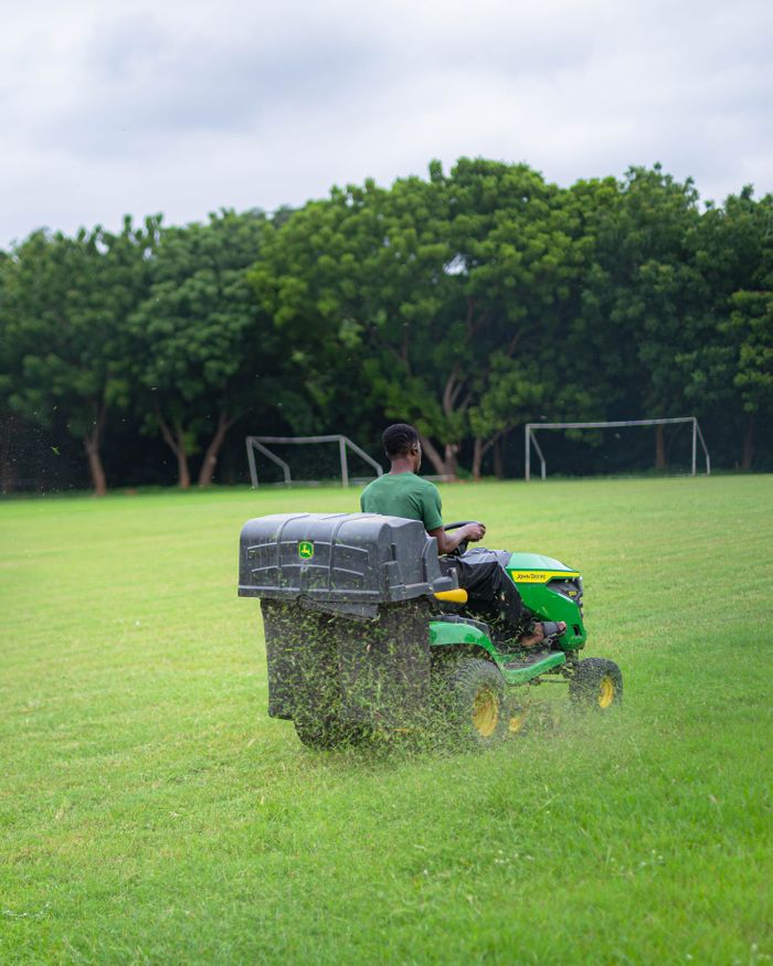 Home Man operating mower on lush sports field in Achimota, Ghana. Greenery and sports goals in view.