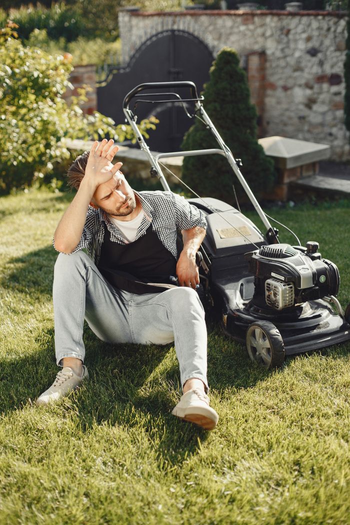About A tired man sitting on the grass with a lawn mower after gardening.